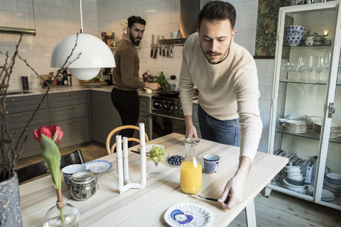 Junger Mann deckt den Tisch, während sein Partner im Hintergrund in der Küche arbeitet, lizenzfreies Stockfoto