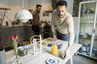 Young man setting table while partner working in background at kitchen - MASF08228