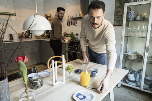 Junger Mann deckt den Tisch, während sein Partner im Hintergrund in der Küche arbeitet - MASF08228