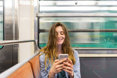 Porträt einer lächelnden jungen Frau in einem U-Bahn-Zug, die auf ihr Smartphone schaut - AFVF00745