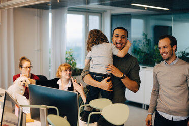 Cheerful businessman carrying son while standing amidst colleagues at creative office - MASF08256