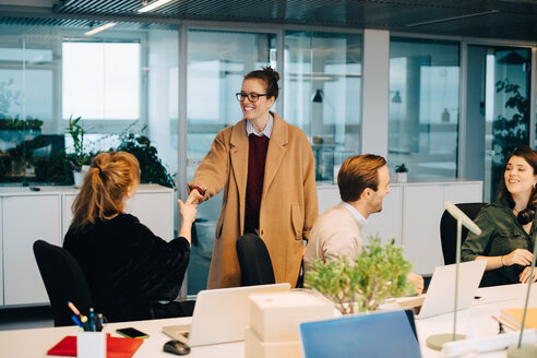 Lächelnde Geschäftsfrau, die eine weibliche Fachkraft begrüßt, die neben Kollegen in einem kreativen Büro sitzt - MASF08259