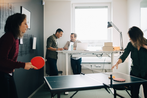 Weibliche Fachkräfte spielen Tischtennis, während männliche Kollegen im Kreativbüro einen Laptop benutzen, lizenzfreies Stockfoto