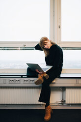 Selbstbewusste junge Geschäftsfrau in voller Länge, die einen Laptop benutzt, während sie auf der Fensterbank eines kreativen Büros sitzt - MASF08299