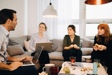 Female professionals looking at businessman sitting with laptop on sofa in creative office - MASF08311