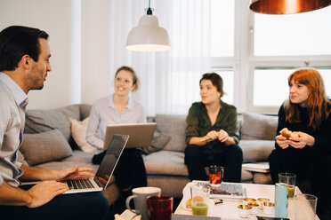 Weibliche Fachkräfte schauen auf einen Geschäftsmann, der mit einem Laptop auf einem Sofa in einem kreativen Büro sitzt - MASF08311