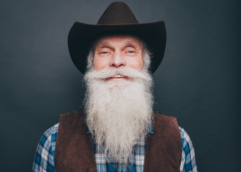 Porträt eines glücklichen, bärtigen älteren Mannes mit Cowboyhut auf grauem Hintergrund, lizenzfreies Stockfoto