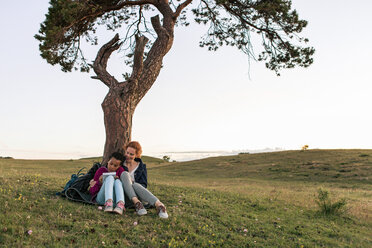 Full length of mother and daughter sitting by tree on grass against sky during sunset - MASF08421