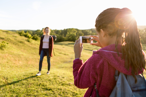 Rückansicht eines Mädchens, das seine auf einem Feld stehende Mutter fotografiert, gegen einen klaren Himmel bei Sonnenuntergang, lizenzfreies Stockfoto