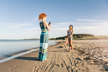 Woman photographing daughter standing on shore at beach against sky - MASF08443