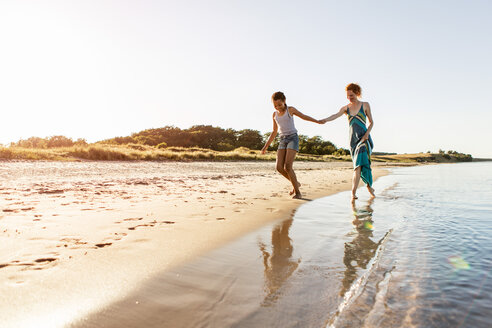 Fröhliches Mädchen in voller Länge, das mit seiner Mutter Händchen hält, während es am Strand gegen den Himmel läuft - MASF08447
