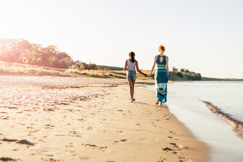 Rückansicht eines Mädchens in voller Länge, das mit seiner Mutter am Strand spazieren geht und dabei die Hände hält, lizenzfreies Stockfoto