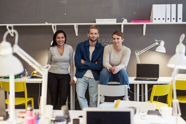 Portrait of confident multi-ethnic business colleagues sitting on desk against wall at creative office - MASF08458