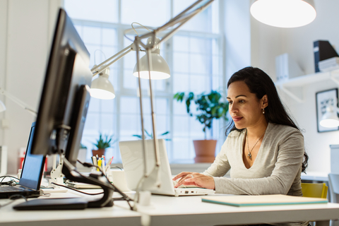 Selbstbewusste Geschäftsfrau im mittleren Erwachsenenalter, die einen Laptop benutzt, während sie in einem kreativen Büro am Schreibtisch sitzt, lizenzfreies Stockfoto