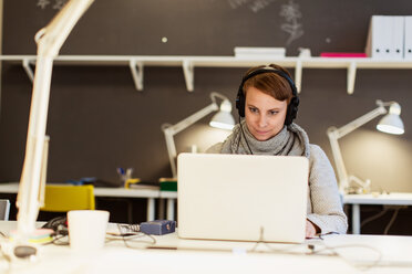 Selbstbewusste Geschäftsfrau hört Kopfhörer, während sie einen Laptop im Kreativbüro benutzt - MASF08468