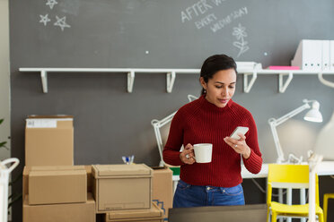 Geschäftsfrau, die eine Kaffeetasse hält, während sie ihr Smartphone an der Wand im Kreativbüro benutzt - MASF08472