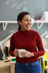 Cheerful businesswoman holding coffee cup while looking away against wall at creative office - MASF08475