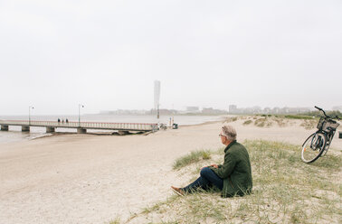 Seitenansicht eines älteren männlichen Pendlers, der im Sand am Strand sitzt, gegen den klaren Himmel - MASF08516