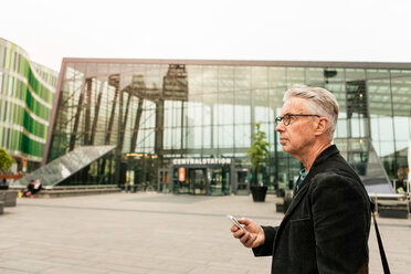 Side view of senior male commuter looking away while holding smart phone against railroad station building - MASF08522