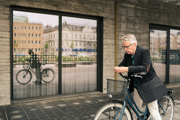 Älterer Mann benutzt sein Smartphone, während er mit dem Fahrrad vor einem Gebäude in der Stadt steht - MASF08523