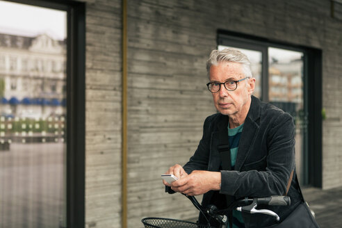 Porträt eines älteren Mannes, der sein Smartphone benutzt, während er mit dem Fahrrad in der Stadt steht - MASF08524