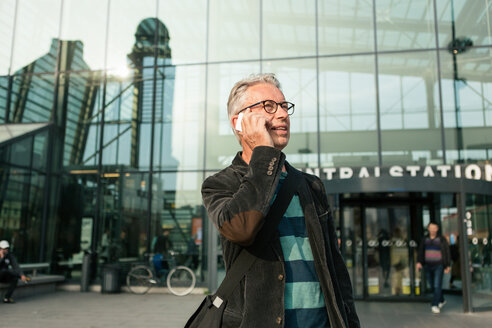 Älterer männlicher Pendler, der wegschaut, während er mit seinem Smartphone am Bahnhofsgebäude spricht - MASF08526