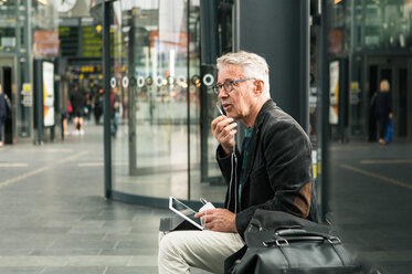 Älterer männlicher Pendler, der sich über In-Ear-Kopfhörer unterhält, während er auf dem Bahnhof bei Taschen sitzt - MASF08529
