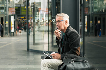 Senior male commuter talking through in-ear headphones while sitting by bags at railroad station - MASF08529