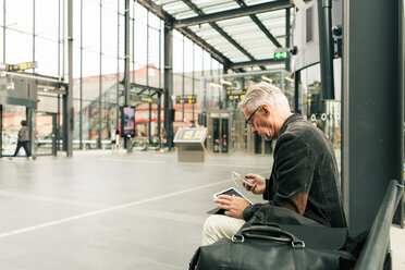 Älterer männlicher Pendler sitzt mit Technik und Taschen am Bahnhof - MASF08533