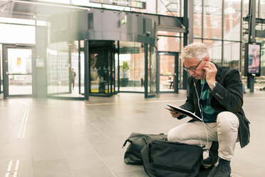 Älterer männlicher Pendler in voller Länge, der ein digitales Tablet in der Hand hält, während er auf dem Bahnhof vor Taschen kauert - MASF08534