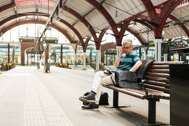 Älterer Mann in voller Länge bei der Nutzung von Technologien, während er auf einer Bank am Bahnhof sitzt - MASF08538
