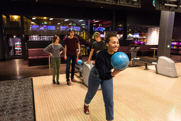 Lächelndes Teenager-Mädchen, das eine Kugel gegen Freunde hält, die auf beleuchtetem Parkett in einer Bowlingbahn stehen - MASF08580