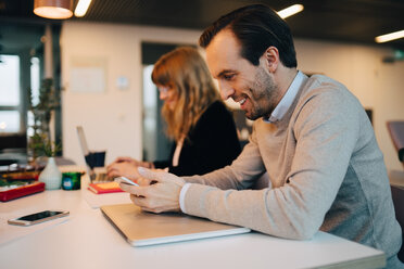 Side view of smiling businessman using smart phone while sitting by businesswoman at desk in creative office - MASF08592