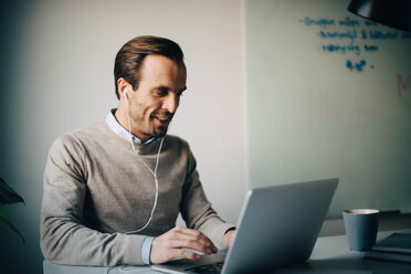 Smiling businessman using laptop while listening to in-ear headphones at creative office - MASF08598