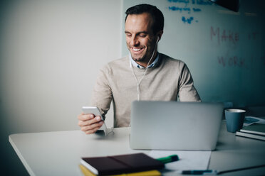 Smiling businessman using smart phone and laptop while sitting at desk in creative office - MASF08599