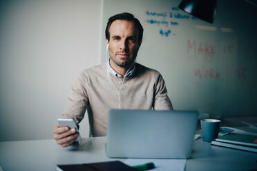 Porträt eines selbstbewussten Geschäftsmannes, der mit Smartphone und Laptop am Schreibtisch in einem kreativen Büro sitzt - MASF08600