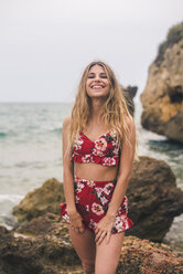 Porträt einer glücklichen jungen Frau, die an einem Felsen am Meer steht - ACPF00098