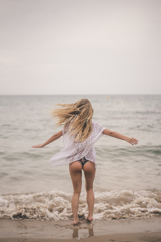 Rückansicht einer jungen Frau, die mit ausgestreckten Armen am Strand steht, lizenzfreies Stockfoto