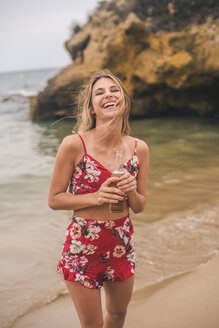 Porträt einer glücklichen jungen Frau, die mit einem Bier am Strand steht - ACPF00102