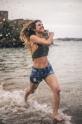 Glückliche junge Frau läuft am Strand und spritzt mit Wasser, lizenzfreies Stockfoto