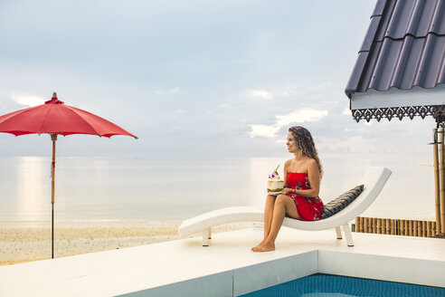 Thailand, Koh Phangan, glückliche Frau sitzt am Pool in der Nähe des Strandes und schaut in die Ferne - MOMF00474