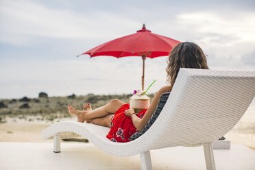 Thailand, Rückenansicht einer Frau, die auf einer Sonnenliege mit einer frischen Kokosnuss sitzt und auf den Strand schaut - MOMF00475