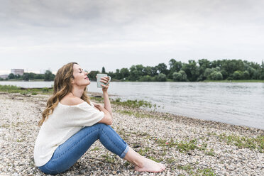 Frau sitzt am Flussufer und genießt eine Tasse Kaffee - UUF14451