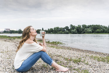 Woman sitting at the riverside enjoying cup of coffee - UUF14451