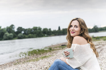 Smiling woman with cup of coffee sitting at the riverside - UUF14452