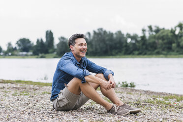 Laughing man sitting at the riverside looking sideways - UUF14458