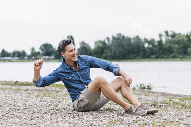 Laughing man sitting at the riverside throwing stones - UUF14459