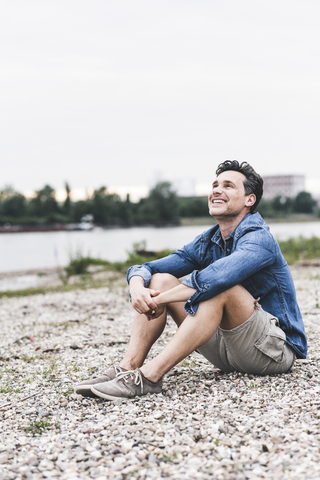 Lächelnder Mann, der am Flussufer sitzt und nach oben schaut, lizenzfreies Stockfoto