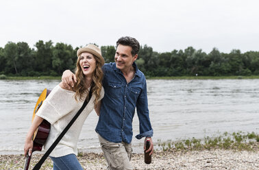 Glückliches Paar, das am Flussufer mit Bierflasche und Gitarre spazieren geht - UUF14495