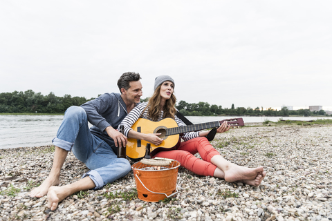 Glückliches Paar mit Bierflasche, Gitarre und Grill, das sich am Flussufer entspannt, lizenzfreies Stockfoto